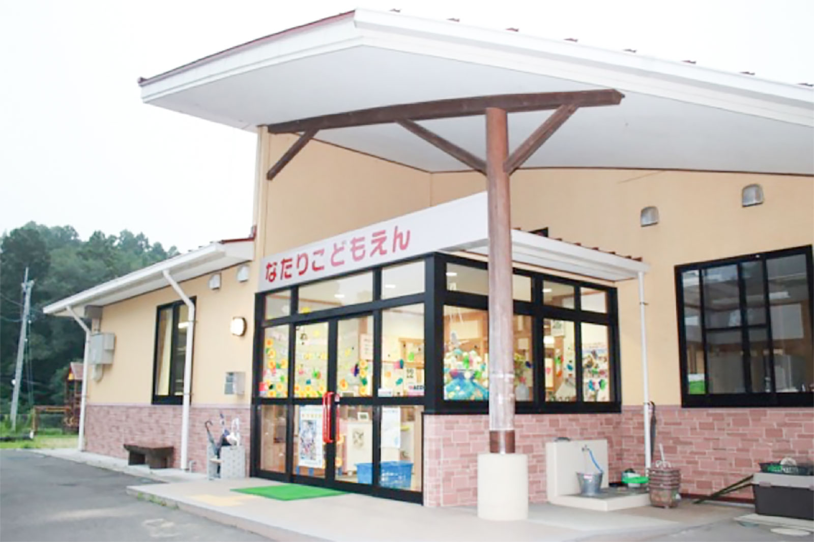 「なたりこどもえん」と書かれた看板が掲げられた、ガラス張りの入り口とベージュとレンガ調の外壁が特徴的な幼児施設の建物外観の写真