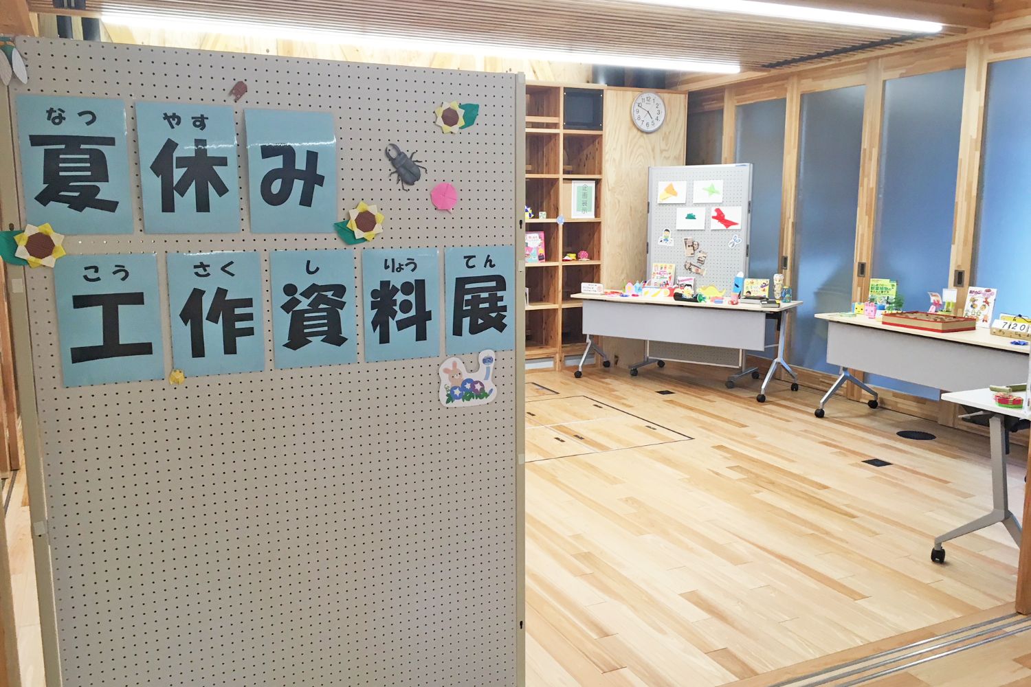 木の壁と床に囲まれた図書館の一角で「夏休み工作資料展」と書かれた展示ボードとテーブルに作品が並ぶ展示コーナーの写真