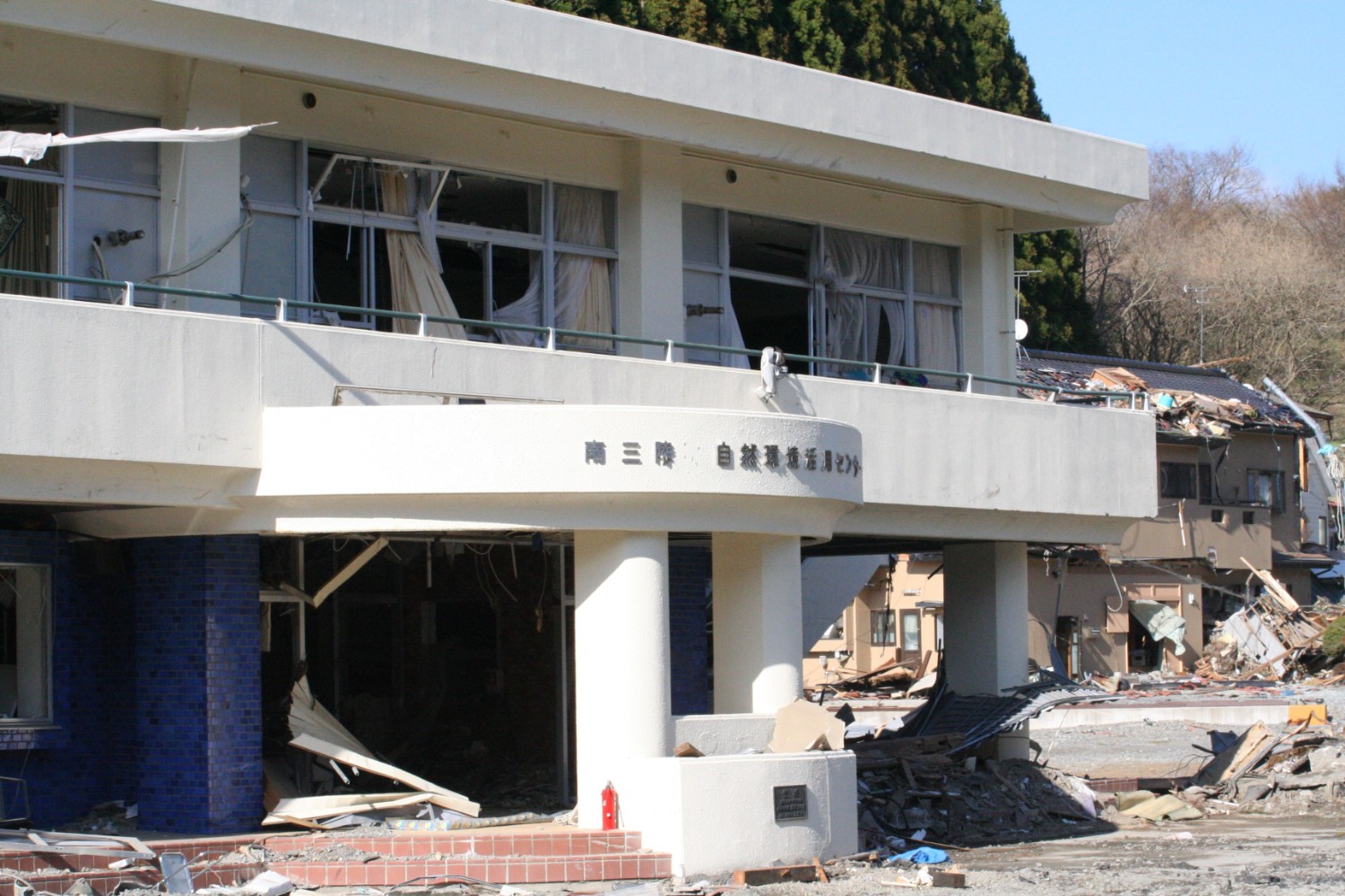 地震や津波の被害により窓ガラスが割れ、壁が崩れ落ちている白い建物と、その周囲に倒壊した家屋や瓦礫が散乱している様子が写っている写真
