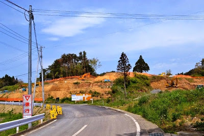 地面が掘り起こされ、むきだしになった土の斜面が整地されており、左へカーブする道路沿いに黄色い注意標識と徐行の看板が立っている工事中の丘の写真