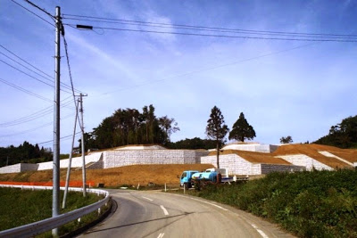 白いコンクリート擁壁が斜面に複数段積まれており、道路脇に水色の工事車両が停まっている造成工事現場の写真