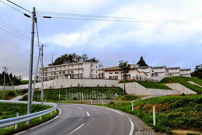 白いコンクリート擁壁の上に住宅が建ち、左に向かってカーブしている道路の先に建つ町営住宅の写真