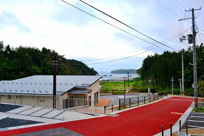 奥に緑の田畑と海が広がっている手前の傾斜地に、赤い舗装道路とフェンスのある建物の屋根が見える写真