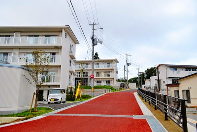 赤い舗装の坂道の両側に白い集合住宅と一戸建てが建ち、左側には駐車された軽自動車と小さな木が立っている町営名足復興住宅の街並みの写真