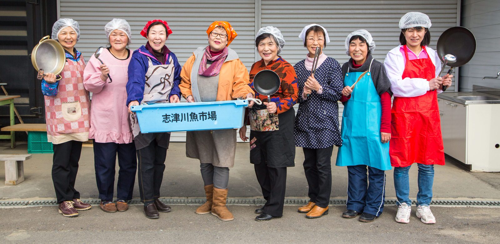 工場の前で、エプロンや三角巾、ヘアーネットを着けた女性たちが鍋やお玉などの調理道具、「志津川魚市場」と書かれた青いコンテナを持ち、明るい笑顔で横一列に並んで立っている写真