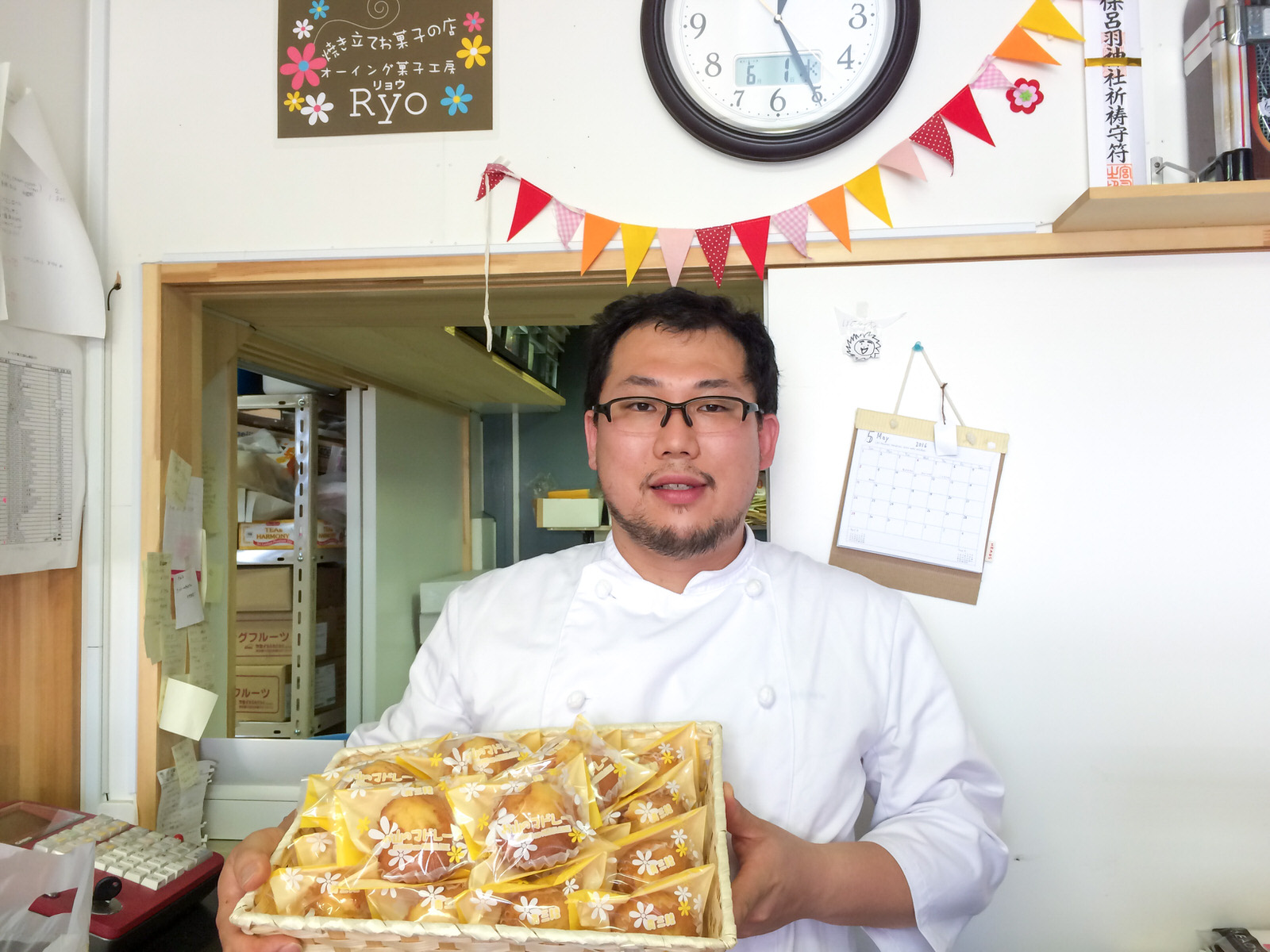 ガーランドなどで装飾された店内で、たくさんの個包装のマドレーヌが入った四角い籠を、コック服を着た男性が抱えている写真