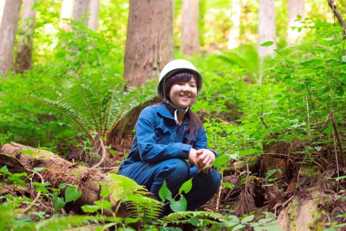 青い作業着と白いヘルメットを身につけた女性が、明るい緑に包まれた森林の中で木に腰かけて笑顔でしゃがんでいる様子の写真