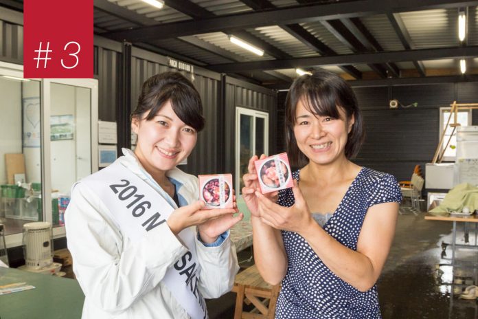 たすきをかけた女性ともう一人の女性が工場の中で笑顔を見せながらホヤの缶詰が入った箱を手に持ち並んで立っている写真の左上に赤い背景に「＃3」と書かれた四角形がある画像