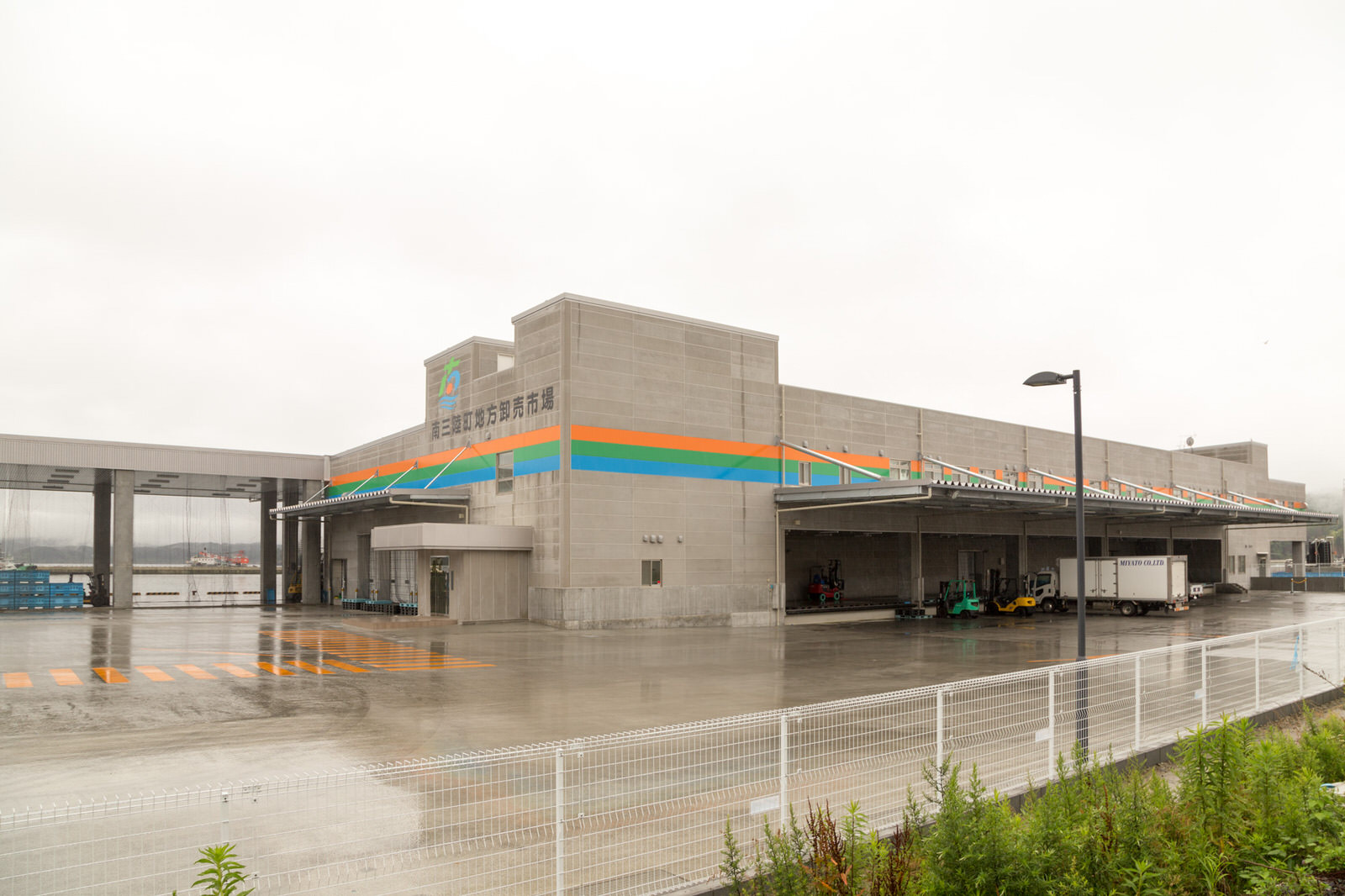 オレンジ、緑、青のラインが入った、南三陸町地方卸売市場の大きな建物が、雨に濡れた広い敷地の中に建っている写真