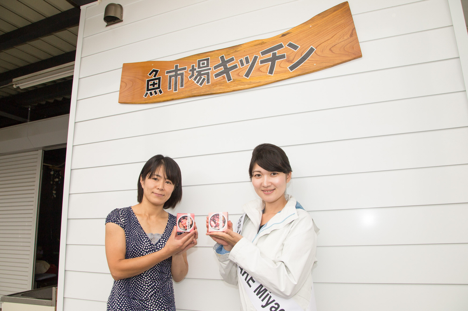 「魚市場キッチン」と書かれた木製の看板が掲げられた白い壁の前で、2人の女性が並んで立ち、手に持った缶詰の箱をカメラに見せている写真