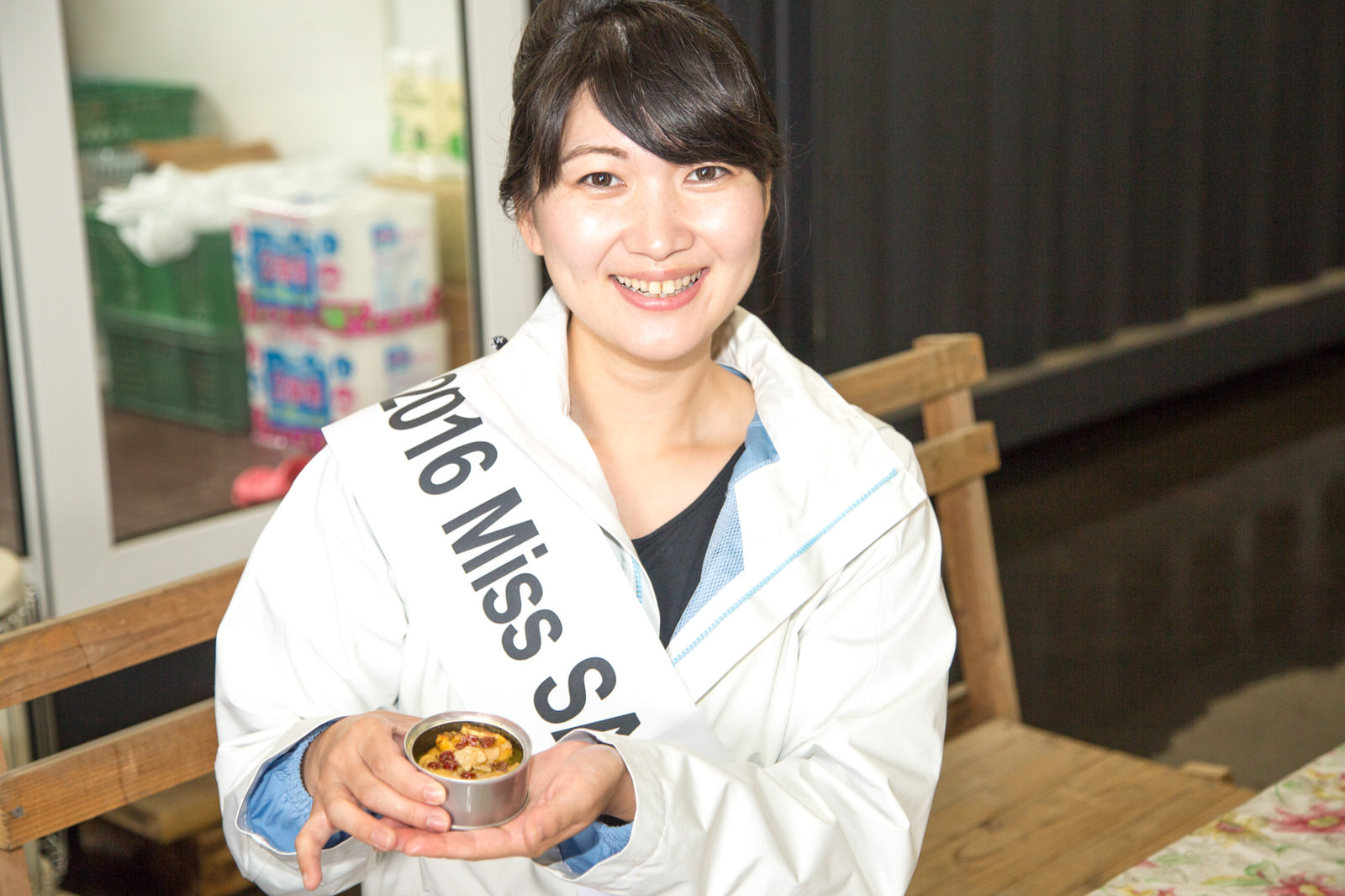 タコのアヒージョの缶詰を持ったタスキをかけた女性が、カメラに笑顔を見せている写真