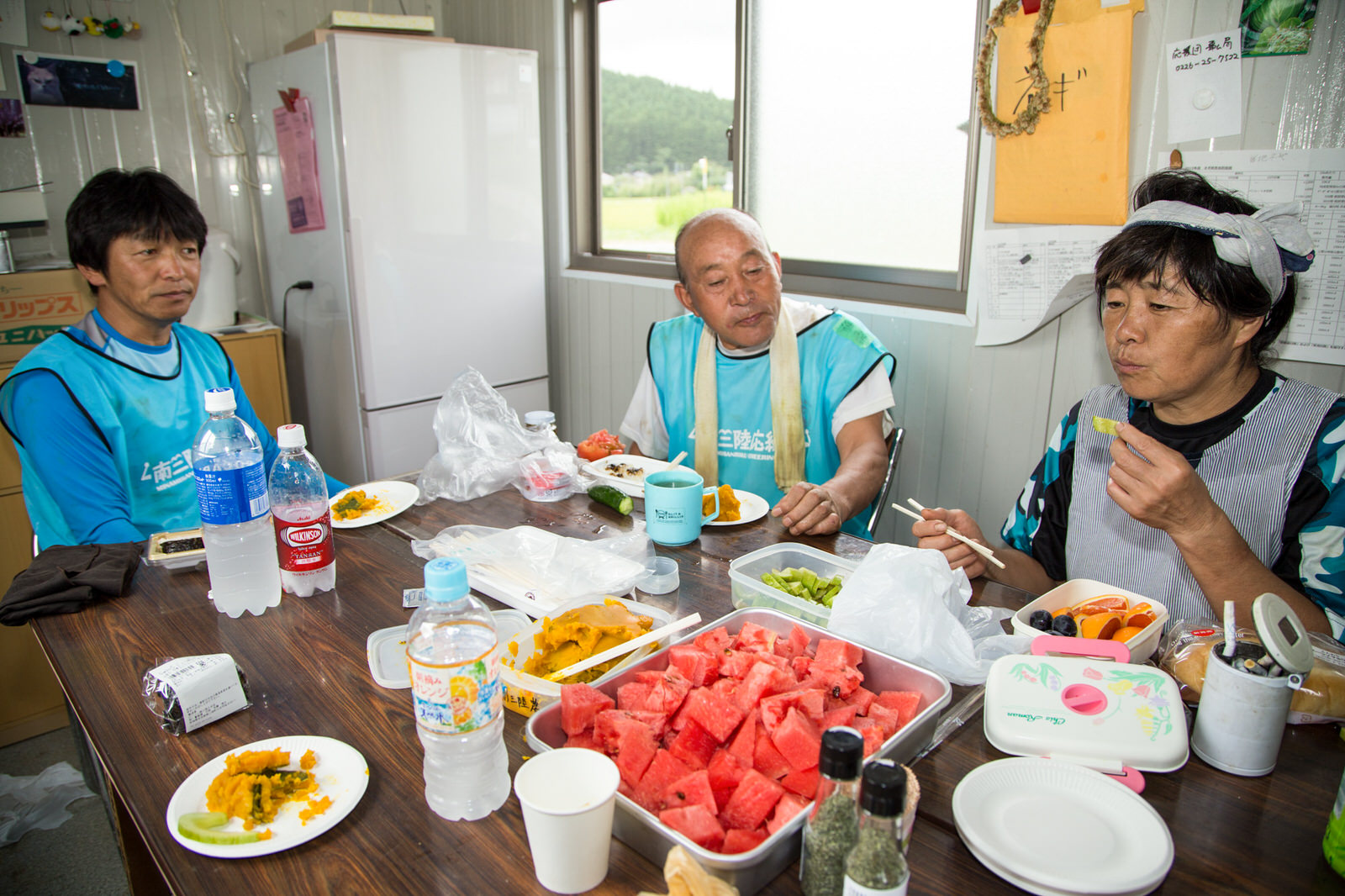 冷蔵庫が設置された室内のテーブルに座った、水色のベストを着た2人の男性と女性スタッフが、カットされたスイカやかぼちゃの煮物などをいただき昼休憩をしている写真
