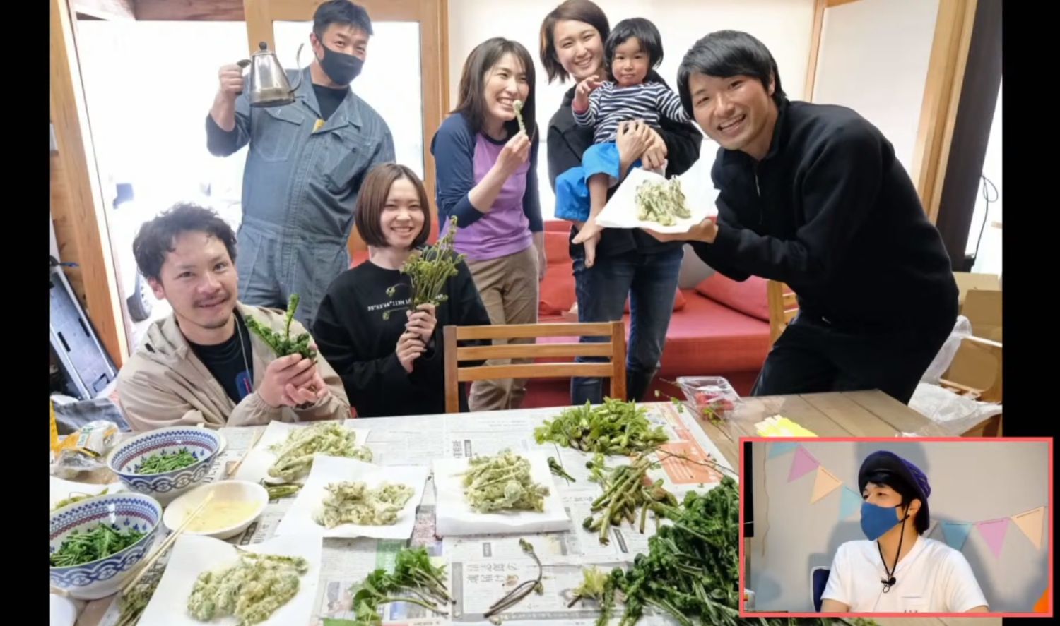 室内のテーブルに山菜の天ぷらや下ごしらえの材料が広げられており、6人の男女と抱っこした子どもが笑顔で山菜を手に持ったり皿を掲げたりしながらポーズをとっており、右下にはその様子を見ている男性のワイプ映像が表示されたスクリーンショット画像