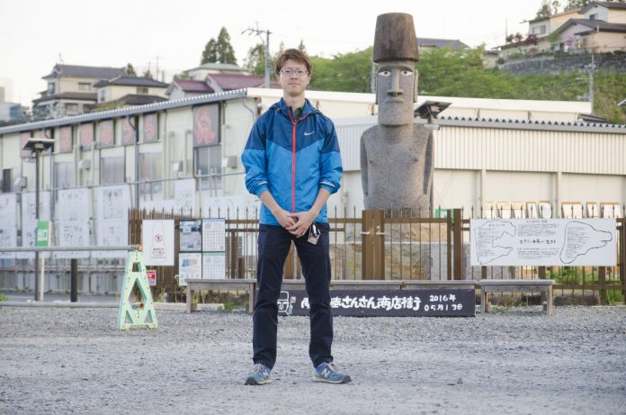 さんさん商店街前のモアイ像を背景に立つ佐藤さんの全身写真