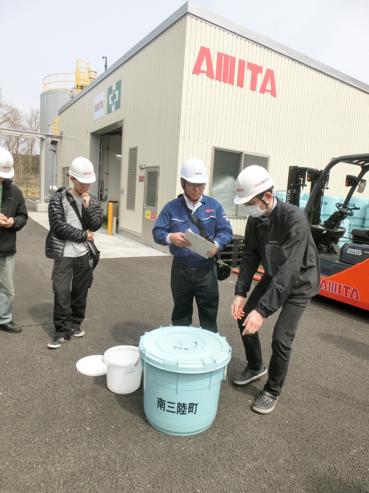 白い壁の四角いAMITAの施設の前で、水色の大きな蓋つきバケツと、小さい白いバケツの周りに、白いヘルメットをかぶった男性が4人集まっている、家庭生ごみの資源化についての説明の際の写真