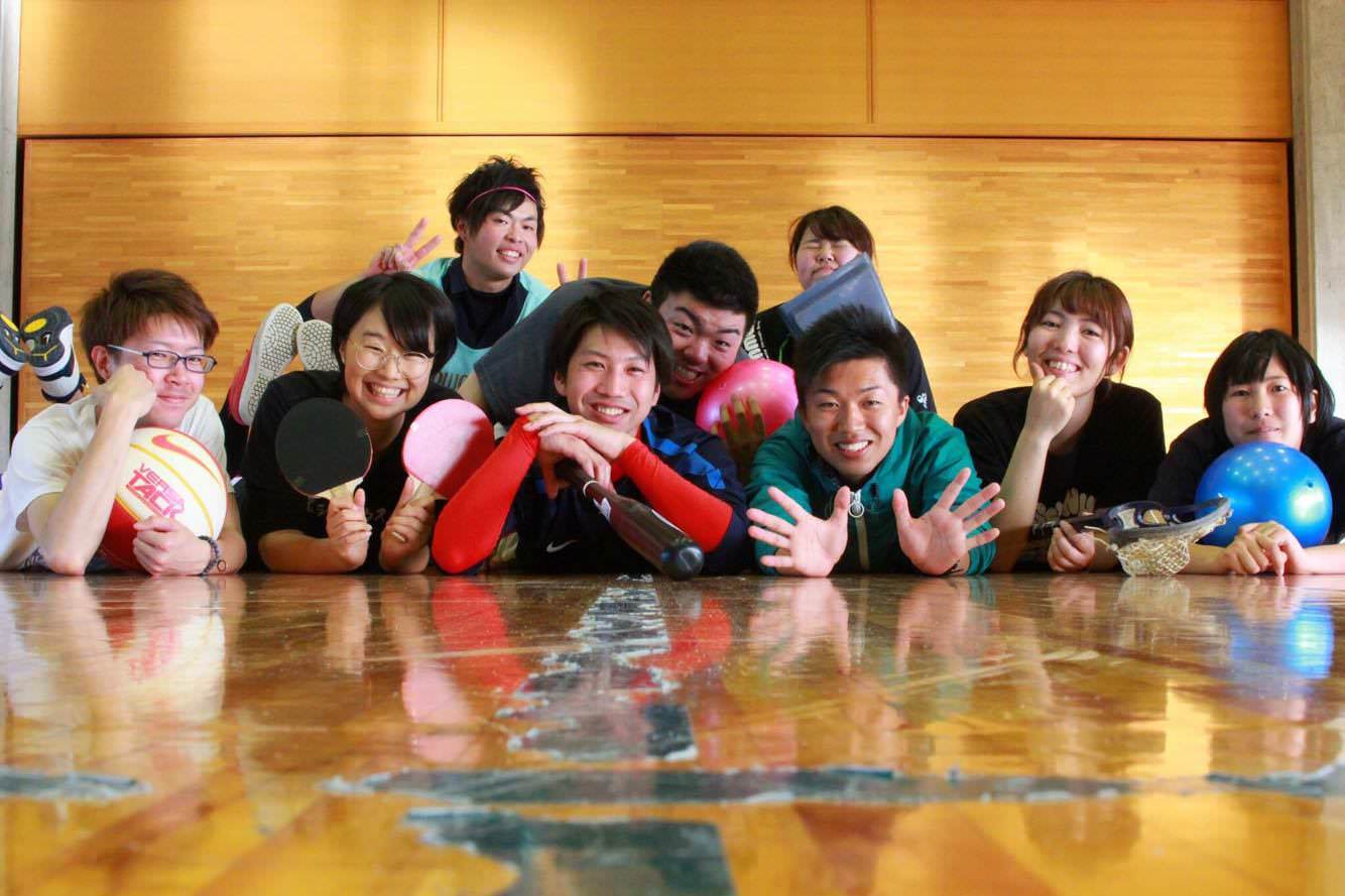 体育館の床に寝そべった佐藤さんと8人の若い男女がバレーボールや卓球ラケットなどを手に笑顔を見せている写真