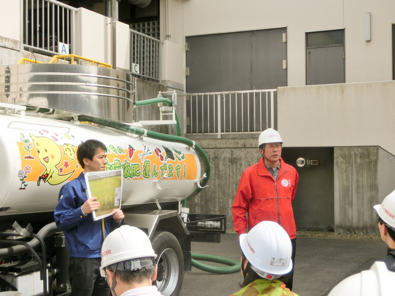 液肥のタンカーの前で、青いジャケットを着た男性が資料を掲げ、隣に立った赤いジャケットを着た男性がほかの参加者の方を見て話をしている写真