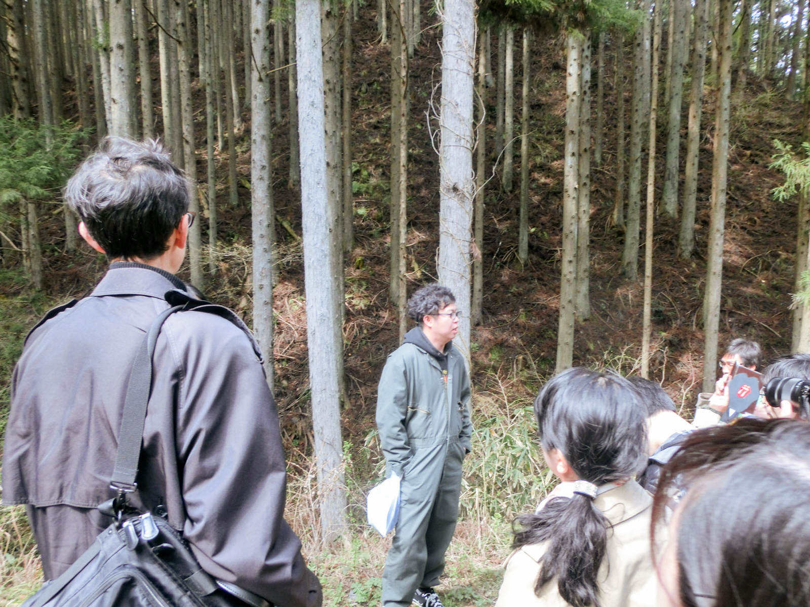間伐された森を背に緑色のつなぎを着た男性が集まった参加者に向かって話をしている写真