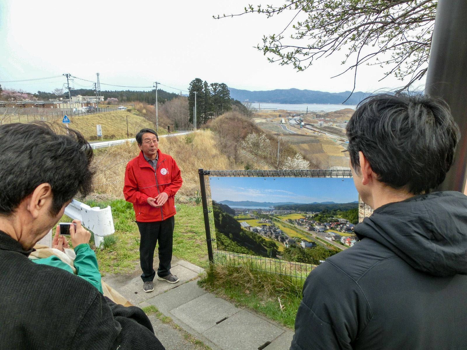 緑に囲まれた水田や住宅の向こうに海が見える震災前の写真が飾られたフェンスの横に赤いジャケットを着た男性が立ち、その背後には茶色い工事現場となった写真と同じ風景が広がっている写真