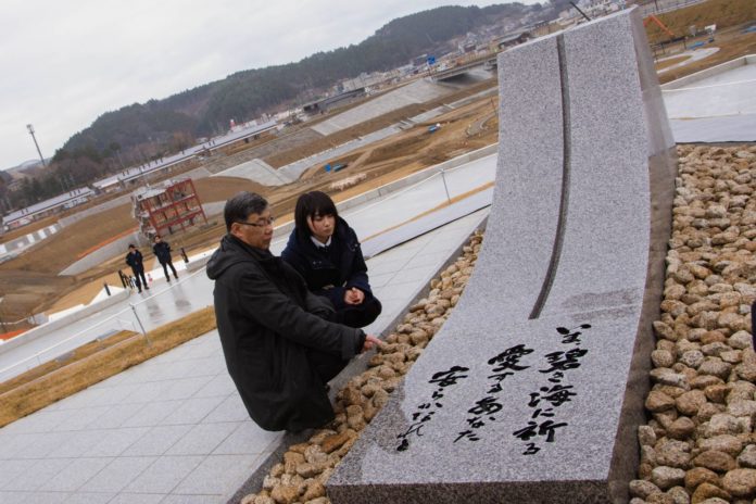 「いま、碧き海に祈る 愛するあなた 安らかなれと」と言葉が刻まれた慰霊碑の前で、男性と女性がが黙祷や祈りを捧げている様子の写真