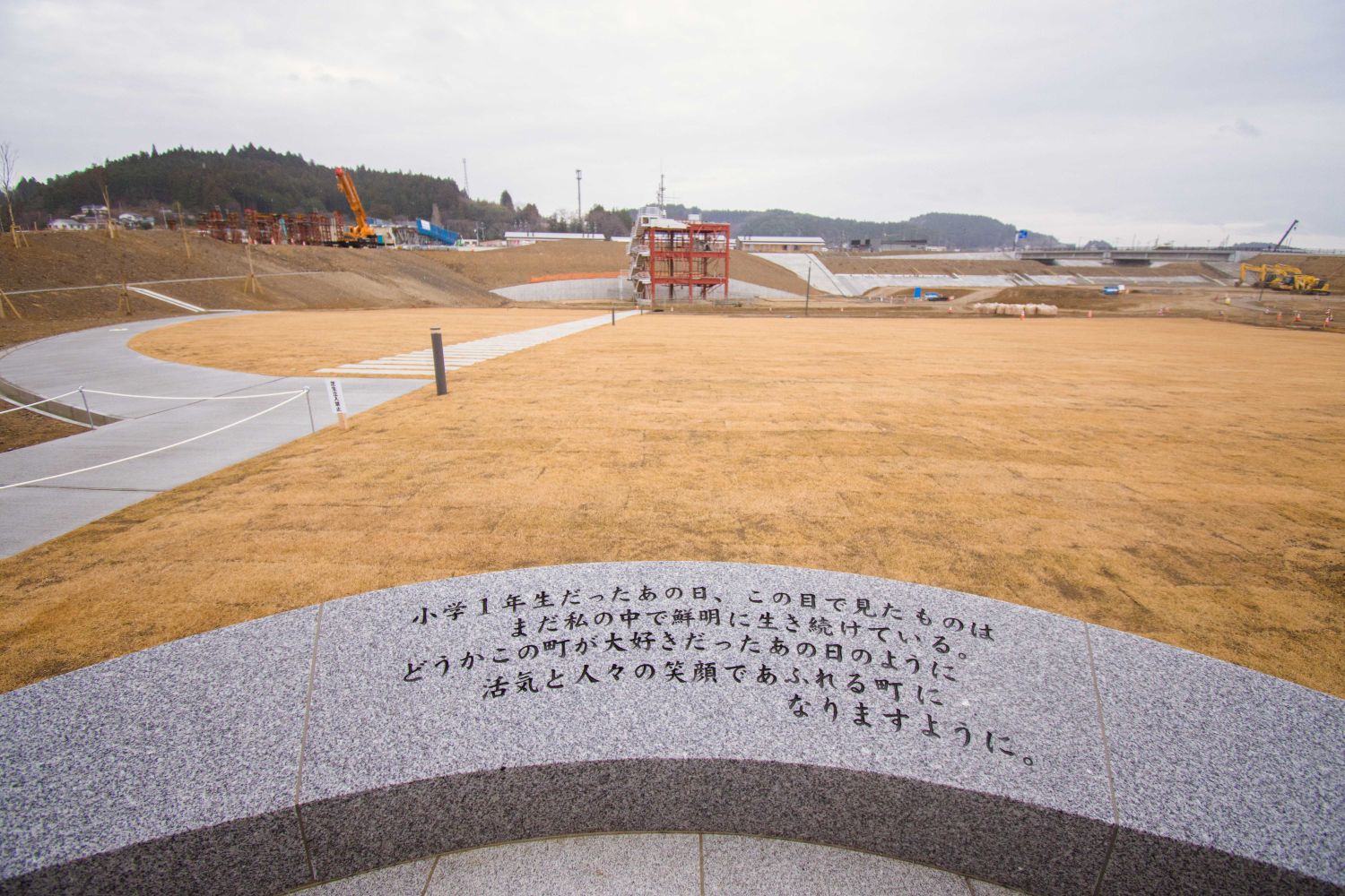 円形の石製に「小学1年生だったあの日、この目で見たものは まだ私の中で鮮明に生き続けている。どうかこの町が大好きだったあの日のように、活気と人々の笑顔であふれる町になりますように。」黒い文字でメッセージが刻まれ、奥に東日本大震災の被災地で整備が進められている様子の写真