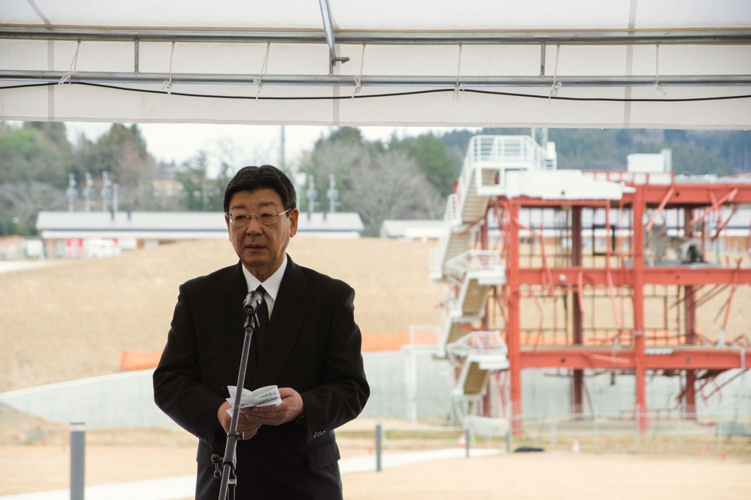 津波の被害を受けた建物を背景に、黒いスーツを着た町長が、マイクの前でスピーチをしている写真