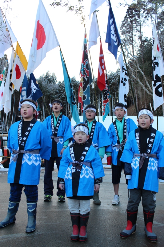 青い法被を着て白い鉢巻きを締めた子どもたちが列になって立ち、背後に日の丸や色とりどりの大漁旗や幟が高く掲げられた屋外で声を合わせている伝統行事「ささよ」の写真