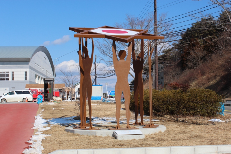 広晴れた青空の下、広場の一角に5体の人影のシルエットが両腕を上げ、中央に日の丸が描かれた天板を掲げるモニュメント「再生と栄光の像」を写した写真