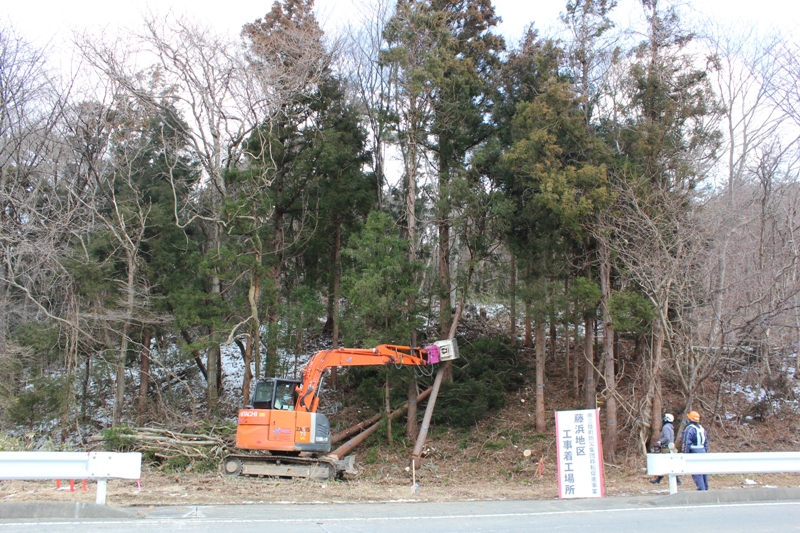 オレンジ色の重機が山の斜面で木を伐採しており、手前には「藤浜地区工事着工場所」と書かれた看板が立ち、作業員2人が作業の様子を見守っている着工式の様子の写真