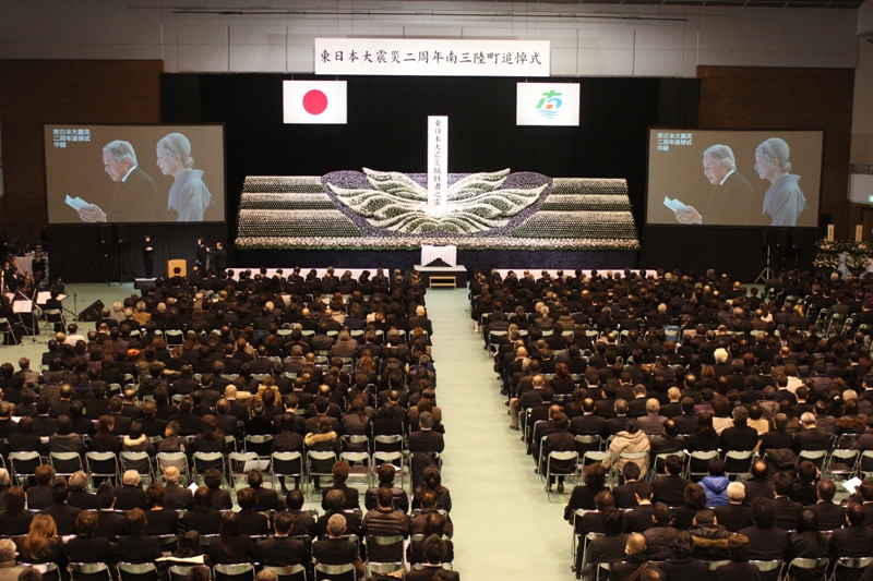 会場いっぱいに参列者が座り、中央の祭壇には白と紫の花で大きな翼のような形が作られ、その中央に「東日本大震災南三陸町犠牲者之霊」と書かれた白い塔が立ち、両脇のスクリーンには天皇皇后両陛下の映像が映し出されている東日本大震災南三陸町追悼式の様子の写真