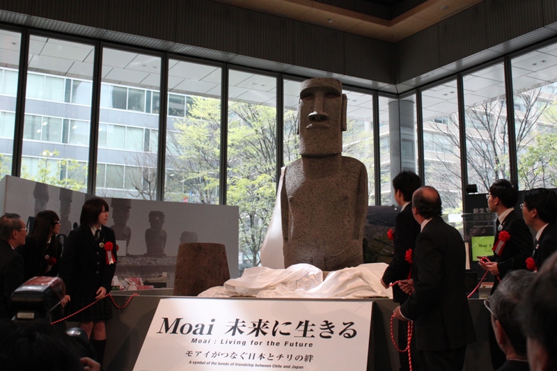 中央に巨大なモアイ像が立ち、手前に「Moai 未来に生きる」と書かれた案内パネルが置かれ、スーツ姿の関係者や女子生徒が像の周りに並んで赤い紐を手にしており、報道カメラや来場者が見守る中で覆いが外されて像が披露されている『Moai 未来に生きる』除幕式の写真