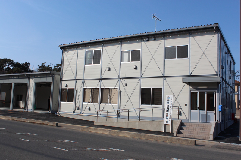 「南三陸消防署仮庁舎」と書かれた看板が玄関前に建てられ、数段の階段とスロープが設置されている2階建ての仮庁舎を写した外観写真