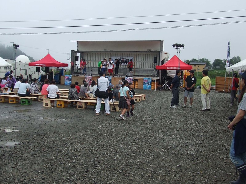 曇り空の下、会場のスレージでバンドの演奏が披露され、観客の方々ベンチに座って楽しんでいる志津川湾夏まつり福興市の会場の様子を写した写真