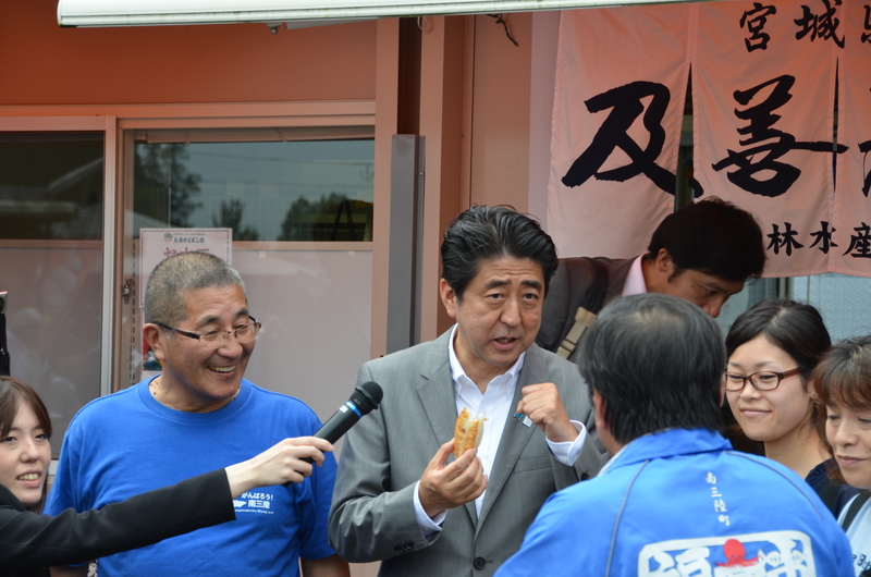 店舗の前で、食べ物を試食している安倍総理がマイクを向けられており、町民の方と話をし、ふれあいをしている様子の写真