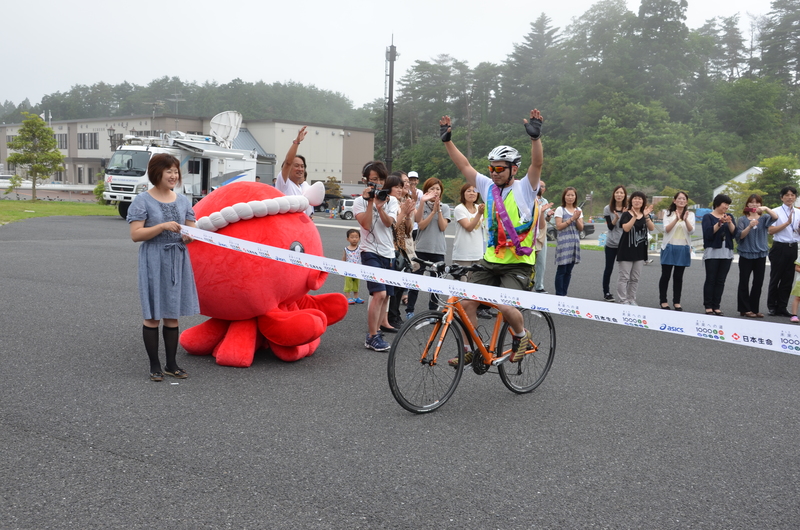 屋外の会場で、自転車に乗ったランナーが両手を挙げてゴールのテープを切ろうとしており、後方ではオクトパス君や多くの人たちが拍手で迎えているリレーのゴールの様子を写した写真