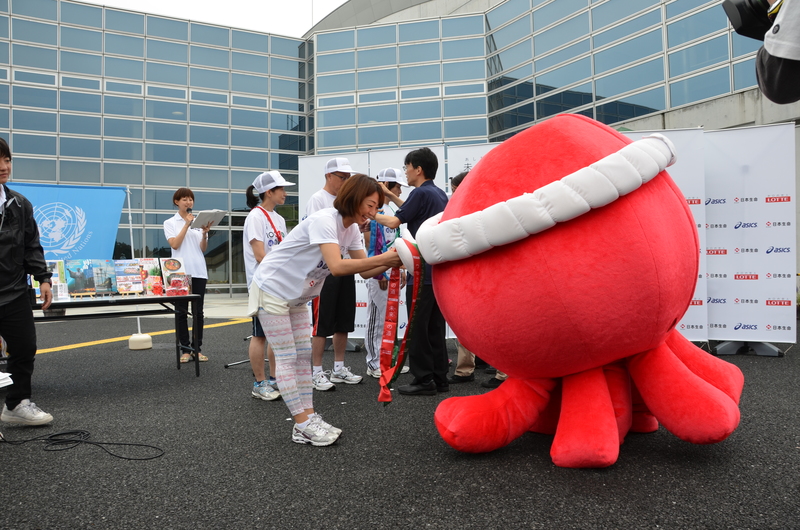 屋外の会場で、オクトパス君から関係者の女性がタスキを受け取っている場面を写した写真