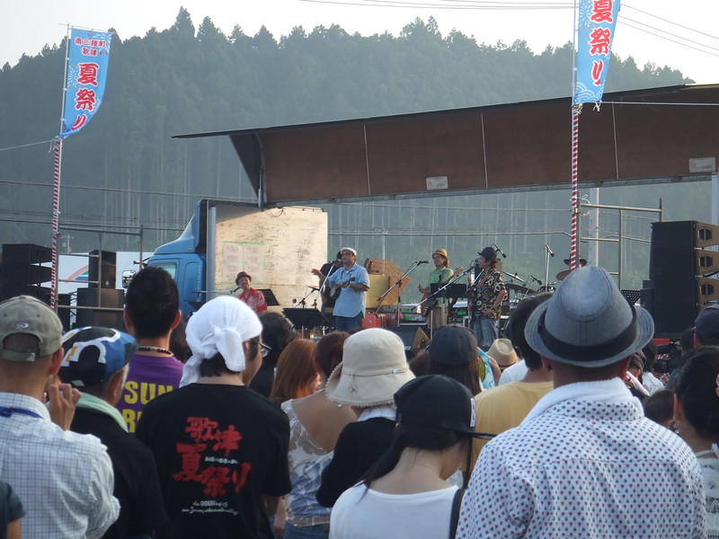 歌津夏まつりの会場で、多くの観客が集まる中、ステージ上でバンドが演奏を披露している歌津夏まつりの様子を写した写真
