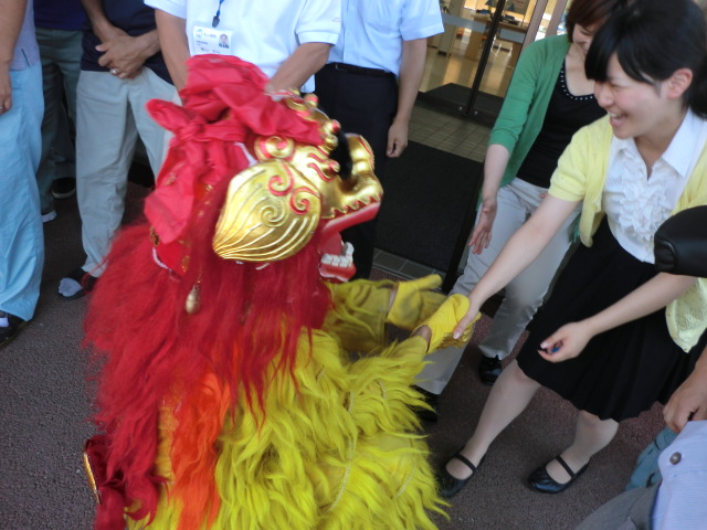 赤と黄色の鮮やかな獅子舞が膝まづき、笑顔の女性と握手を交わしている様子を写した写真