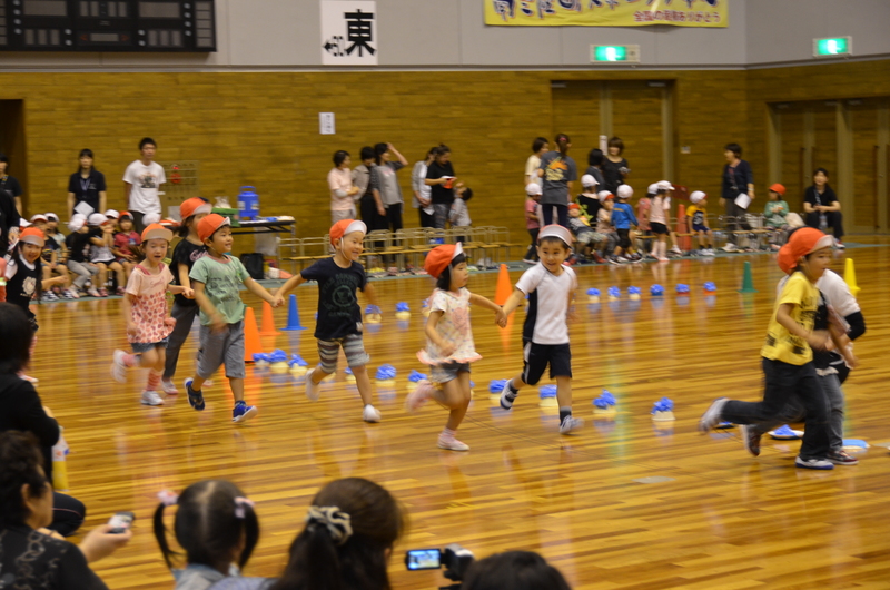 体育館の中で赤い帽子をかぶった園児たちが手をつなぎながら元気に走り、観客席には多くの保護者が見守っている志津川保育所運動会の様子を写した写真