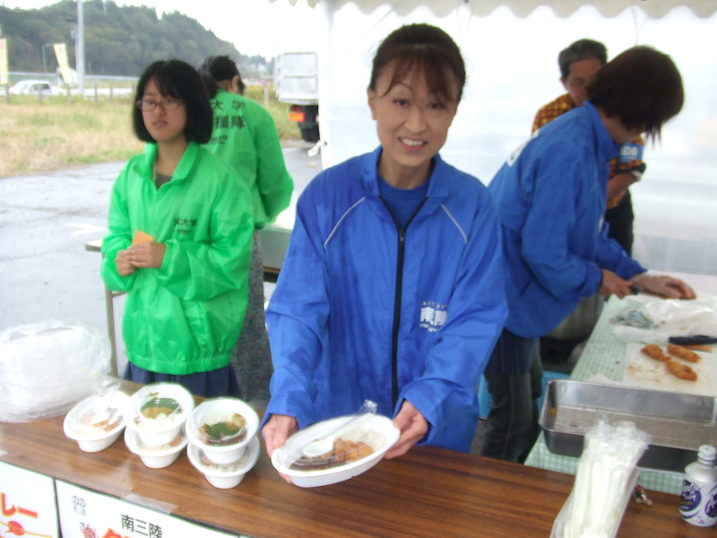 屋外のテントの下で、青いジャンパーを着た女性が笑顔で皿に盛りつけた料理を手に持ち、机の上には他の料理も並べられている、南三陸町のブースを写した写真