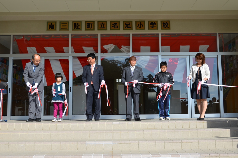 南三陸町立名足小学校の校舎玄関前で、町長と児童、関係者が白い手袋をはめ、赤と白のリボンを持ってテープカットをしている入校式の様子を写した写真