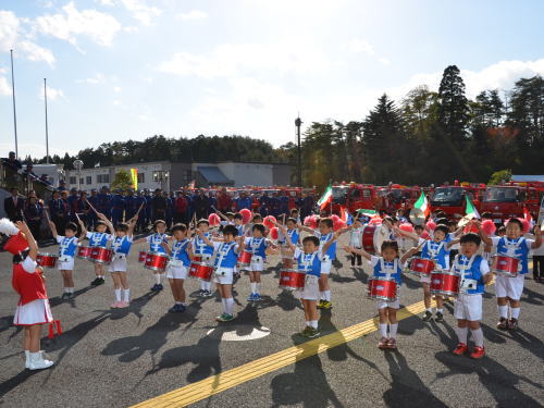 後方に赤い消防車が並んで駐車されている屋外の会場で、赤い衣装の指揮者と青い衣装のあさひ幼稚園鼓笛隊が小太鼓を演奏している防火キャラバン出発式の様子を写した写真