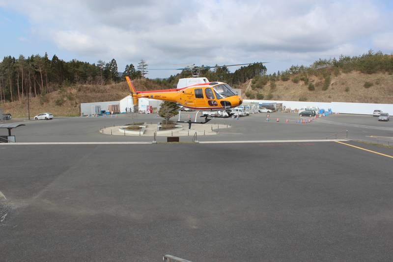 広い駐車場の中央で、NPO法人オールラウンドヘリコプターの黄色いヘリコプターが地面から少し浮かび、離発着訓練を行っている様子の写真
