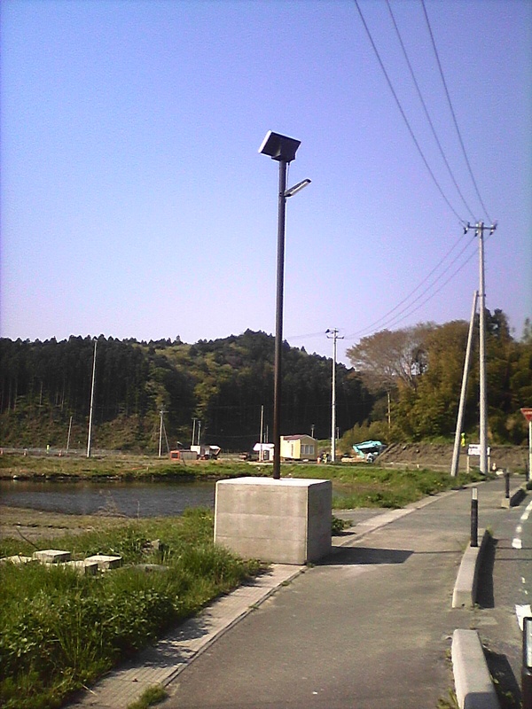 歩道脇に設置されたコンクリートの基台の上に、太陽光パネルを備えたソーラー電灯が立っている様子の写真