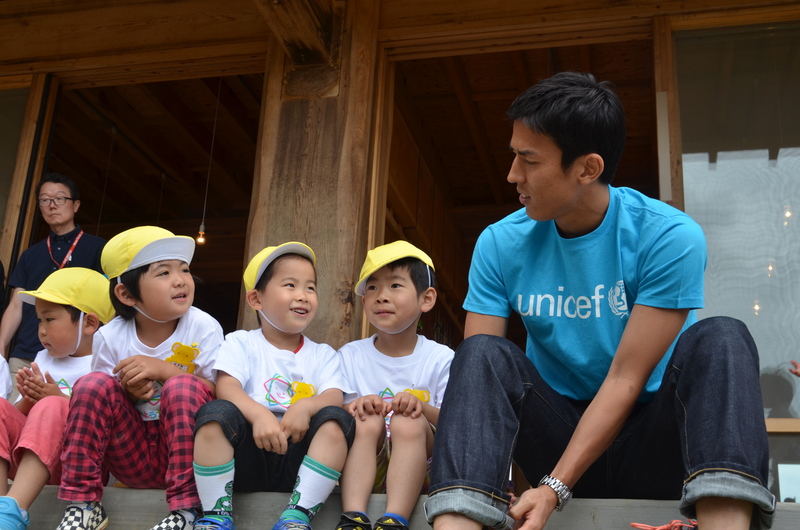 建物の縁側に青いTシャツを着た長谷部誠選手と黄色い帽子をかぶった園児たちが並んで座り、笑顔で会話している様子の写真