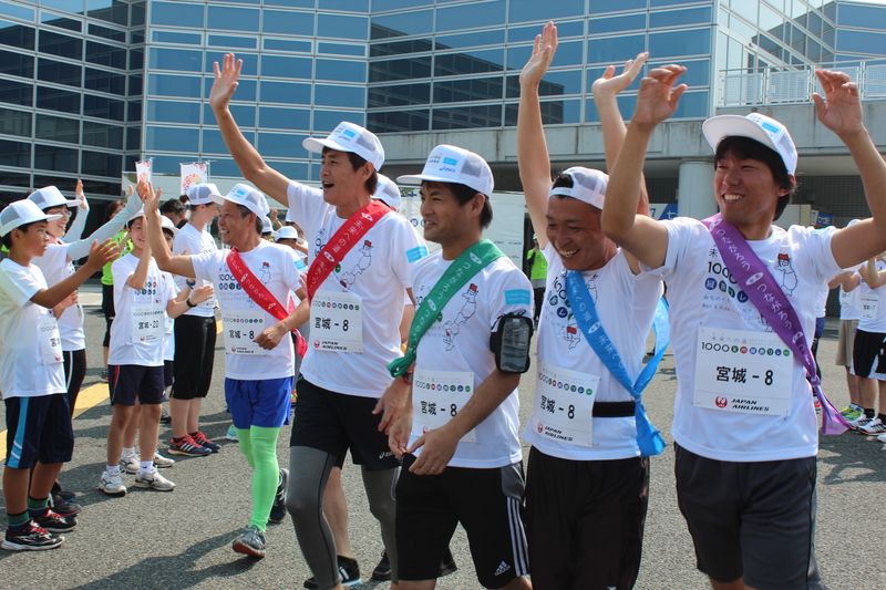 晴天のもと、Tシャツと帽子を身につけた参加者のランナーたちが笑顔で手を上げ、沿道の人々とハイタッチを交わしている未来への道1000km縦断リレー南三陸町出発式の様子の写真