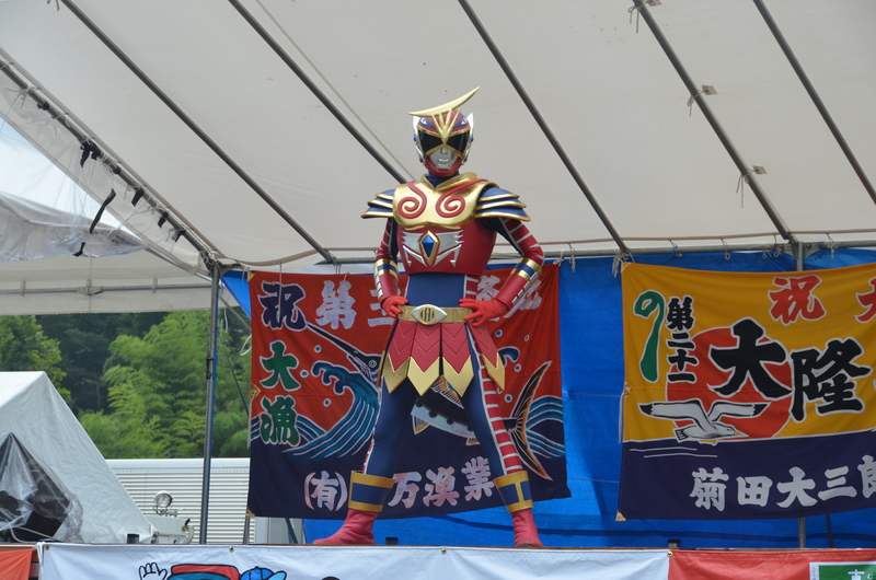 後方に大漁旗が掲示された舞台に赤と金の甲冑風の衣装にマスクを着けたニューヒーロー『未知ノ国守ダッチャー』が両手を腰に当ててポーズを決めて立っている歌津復興夏まつりの様子の写真