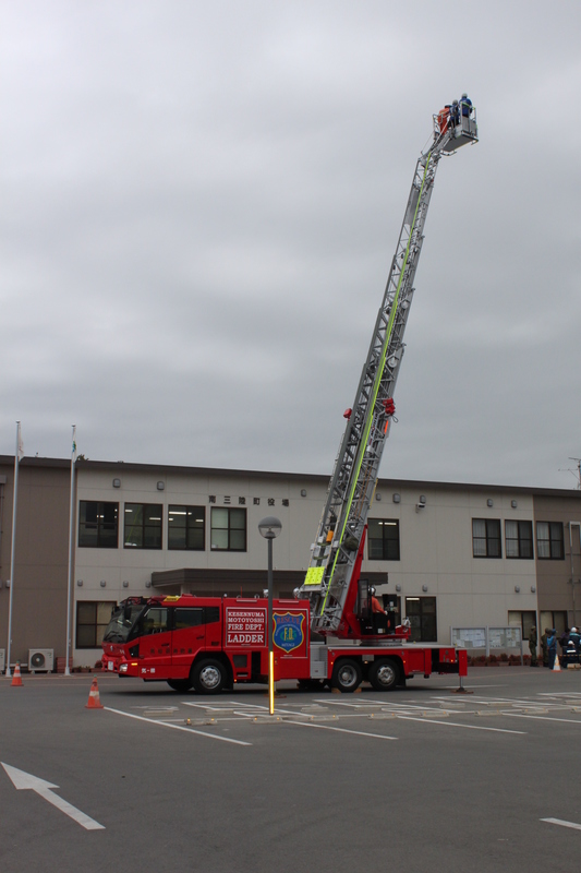 庁舎前の駐車場で行われた総合防災訓練で、赤いはしご車のはしごが高く伸ばされ、搭乗体験が行われている様子の写真
