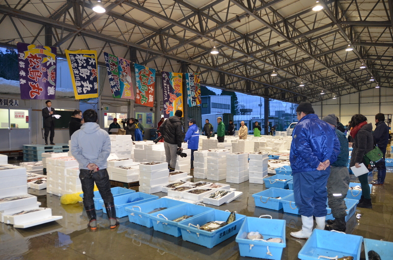 大漁旗が飾られた市場の建物の中で、青いプラスチックの容器や白い発泡スチロール箱に魚介類が並べられ、漁師や仲買人たちが集まっている初セリの様子の写真