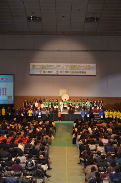 広い屋内会場で、ステージの上には緑や黄色の服を着た多くの人々が並び、客席には大勢の観客が座ってステージを鑑賞している南三陸ボランティア感謝のつどいの写真